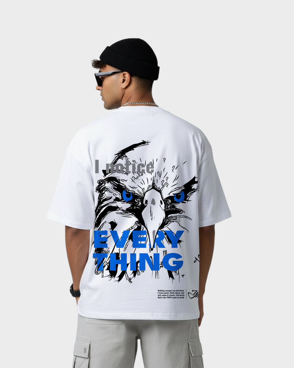 Back view of model wearing TWYN white oversized t-shirt featuring bold eagle illustration with blue eyes and ‘I notice everything’ graphic text
